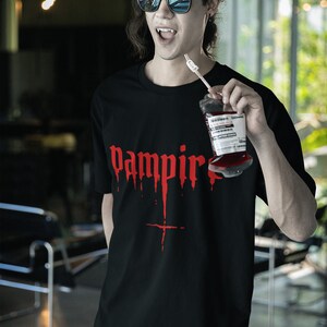May include: A black t-shirt with a red graphic that says "vampire" with dripping blood. The person wearing the shirt is holding a bag of fake blood.