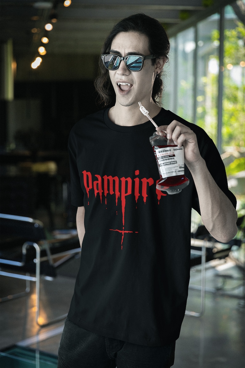 May include: A black t-shirt with a red graphic that says "vampire" with dripping blood. The person wearing the shirt is holding a bag of fake blood.