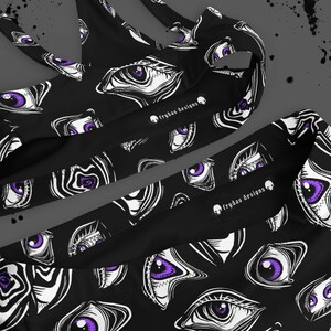 May include: Black athletic top and bottoms set with a repeating pattern of white and purple stylized eyes. The text "frydas designs" is printed on the garment.