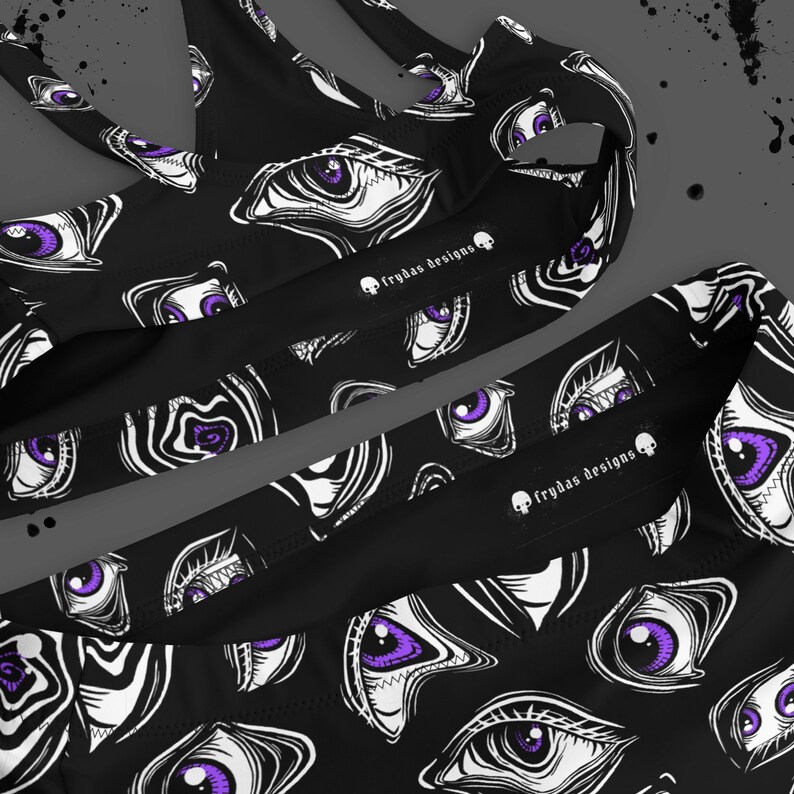 May include: Black athletic top and bottoms set with a repeating pattern of white and purple stylized eyes. The text "frydas designs" is printed on the garment.