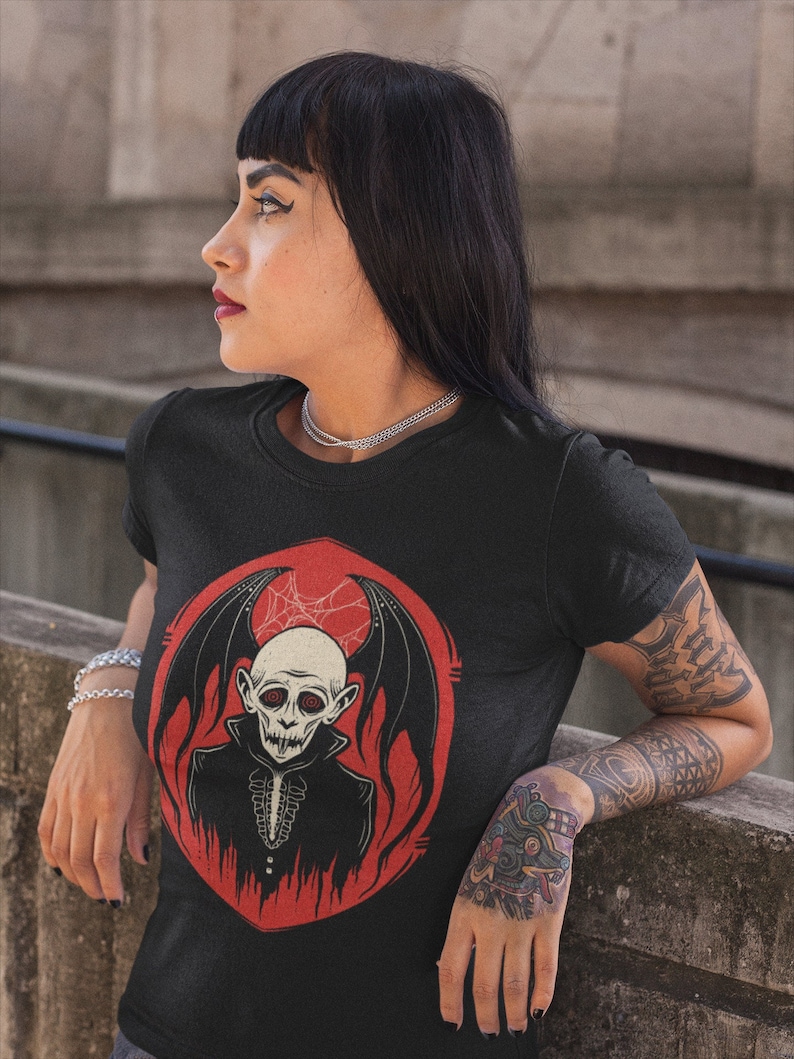 K&ouml;nnte beinhalten: Schwarzes T-Shirt mit einem Aufdruck eines Vampirs mit roten Augen und Fledermausfl&uuml;geln. Der Vampir tr&auml;gt einen schwarzen Anzug und einen wei&szlig;en Kragen. Der Aufdruck ist auf einem roten Hintergrund mit einem Spinnennetz.