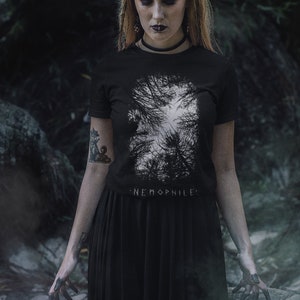 May include: A woman wearing a black t-shirt with a white graphic of trees and the text "NEMOPHILE". She is also wearing a black skirt and has a silver crescent moon on her forehead.