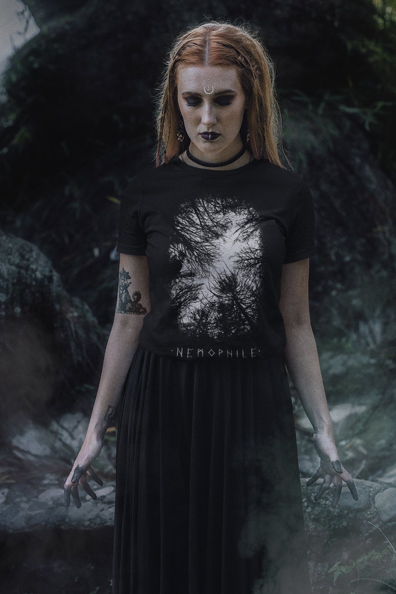 May include: A woman wearing a black t-shirt with a white graphic of trees and the text "NEMOPHILE". She is also wearing a black skirt and has a silver crescent moon on her forehead.