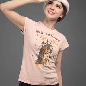 Puede incluir: Una mujer con una camiseta rosa con un gr&aacute;fico de una figura de Krampus y el texto "Gruss vom Krampus". Lleva un gorro de Pap&aacute; Noel y mira hacia arriba.