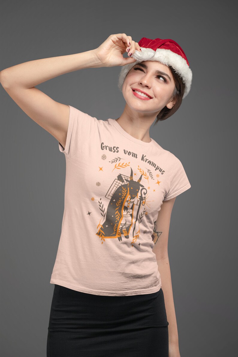 Puede incluir: Una mujer con una camiseta rosa con un gr&aacute;fico de una figura de Krampus y el texto "Gruss vom Krampus". Lleva un gorro de Pap&aacute; Noel y mira hacia arriba.