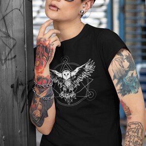 K&ouml;nnte beinhalten: Schwarzes T-Shirt mit einem wei&szlig;en Grafikdruck eines fliegenden Eulen innerhalb eines geometrischen Designs. Die Eule hat einen wei&szlig;en K&ouml;rper und schwarze Augen.