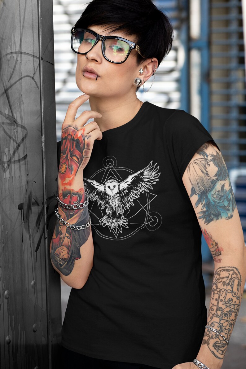 K&ouml;nnte beinhalten: Schwarzes T-Shirt mit einem wei&szlig;en Grafikdruck eines fliegenden Eulen innerhalb eines geometrischen Designs. Die Eule hat einen wei&szlig;en K&ouml;rper und schwarze Augen.