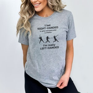 May include: Gray t-shirt with a black graphic that says "I bat RIGHT-HANDED to give everyone else a chance. I'm really LEFT-HANDED". The graphic also includes three baseball players.