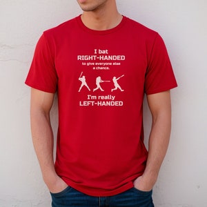 May include: Red t-shirt with a white graphic of a baseball player batting. The text on the shirt reads "I bat RIGHT-HANDED to give everyone else a chance. I'm really LEFT-HANDED."