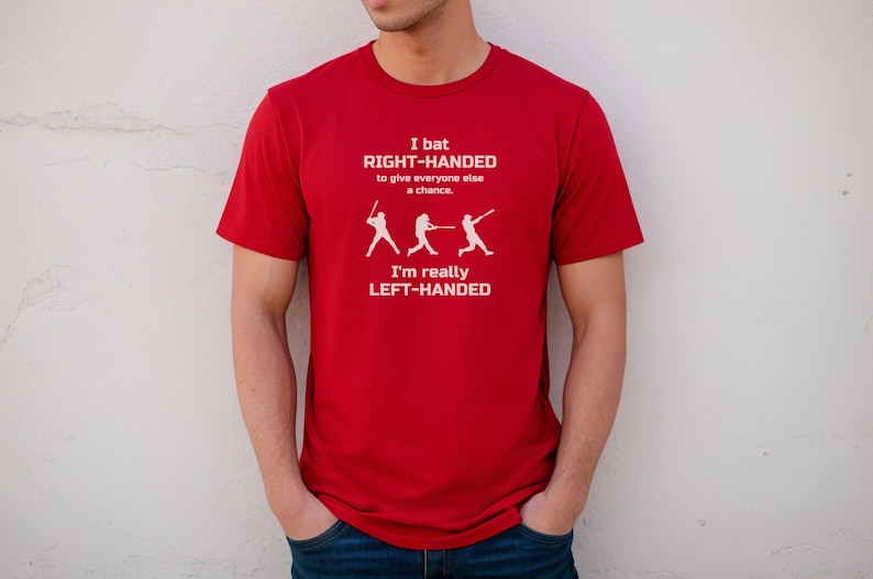 May include: Red t-shirt with a white graphic of a baseball player batting. The text on the shirt reads "I bat RIGHT-HANDED to give everyone else a chance. I'm really LEFT-HANDED."