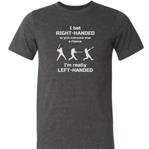 May include: A dark heather grey t-shirt with a white graphic of three baseball players. The text on the shirt reads "I bat RIGHT-HANDED to give everyone else a chance. I'm really LEFT-HANDED."