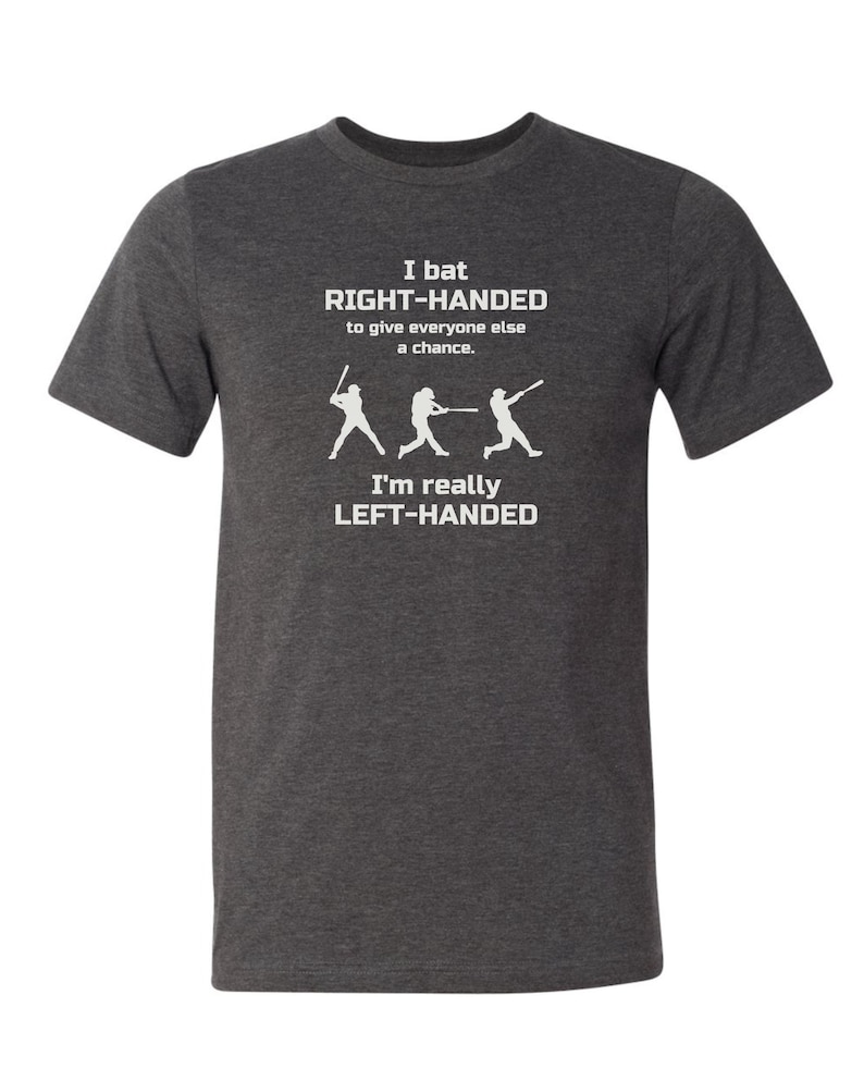 May include: A dark heather grey t-shirt with a white graphic of three baseball players. The text on the shirt reads "I bat RIGHT-HANDED to give everyone else a chance. I'm really LEFT-HANDED."