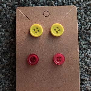 Puede incluir: Pendientes con dos pares de tachuelas en forma de botón. La fila superior tiene tachuelas amarillas con cuatro agujeros, y la fila inferior tiene tachuelas rojas con cuatro agujeros. Los pendientes se presentan en una tarjeta de pendientes marrón.