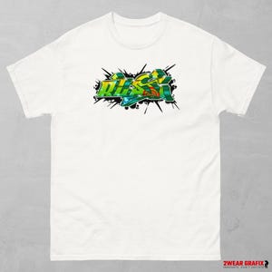 Könnte beinhalten: Weißes T-Shirt mit einem farbenfrohen Graffiti-Design, das "DUSK" in Grün, Gelb und Blau zeigt. Das Design ist von schwarzen Spritzern umgeben. Auf der unteren rechten Ecke des Shirts steht der Text "2WEAR GRAFIX INNOVATE, DON'T IMITATE".