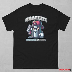 Peut inclure: T-shirt noir avec une illustration de dessin animé d'une bombe de peinture argentée avec un bouchon rouge, tenant une bombe de peinture rose. Le mot "GRAFFITI" est au-dessus de l'illustration. Le t-shirt porte également le texte "EXPRESSION".