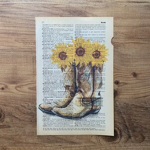 May include: A pair of brown cowboy boots with a distressed white finish, set against a vintage dictionary page with a sunflower illustration. The text on the page includes the word "Arab" and a definition.