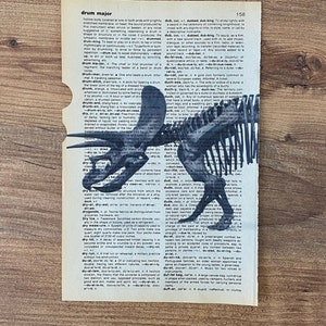 May include: An antique dictionary page with the word "drum major" and a black and white illustration of a dinosaur skeleton. The page is aged with visible wear and tear, and the background is a wooden surface.