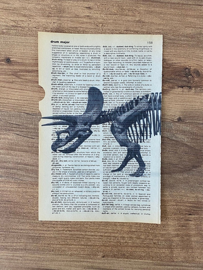 May include: An antique dictionary page with the word "drum major" and a black and white illustration of a dinosaur skeleton. The page is aged with visible wear and tear, and the background is a wooden surface.