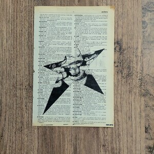 Pu&ograve; includere: Una pagina rettangolare tratta da un vecchio dizionario con carta invecchiata color bianco sporco e testo stampato in nero. Una grande stella nera a cinque punte &egrave; stampata sopra il centro del testo, con un aspetto usurato e strutturato.
