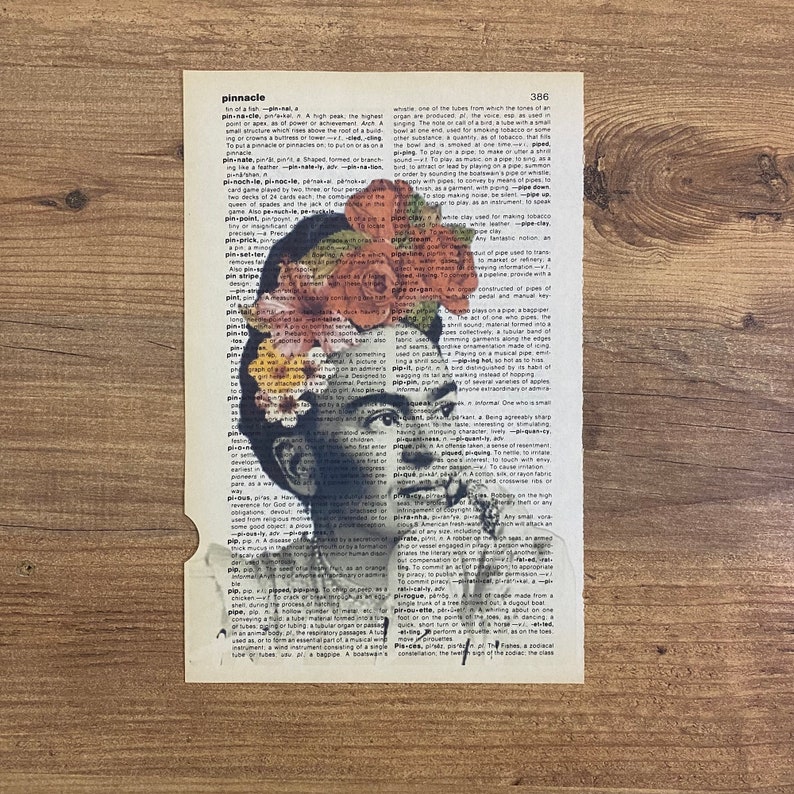 Op de afbeelding: Een zwart-wit portret van Frida Kahlo met bloemen op haar hoofd. Het portret is afgedrukt op een pagina uit een woordenboek met het woord "pinnacle" gemarkeerd.