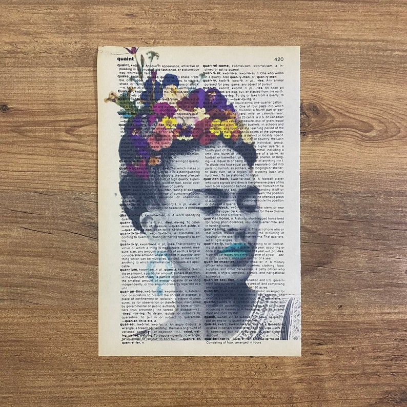Op de afbeelding: Een zwart-wit portret van Frida Kahlo met een kleurrijke bloemenkroon. Het portret is over een woordenboekpagina gelegd met het woord "quaint" dat is gemarkeerd. De afbeelding is een unieke en artistieke weergave van de beroemde Mexicaanse schilder.