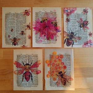May include: Five vintage dictionary pages with watercolor bee illustrations. Each page features a different bee design, with varying colors and styles, including pink and orange accents. The pages are arranged on a wooden surface.