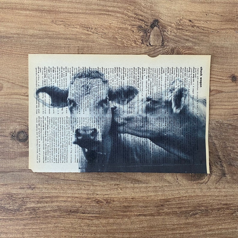 May include: Monochrome cow print on a vintage dictionary page. The artwork depicts two cows in shades of grey, set against a background of printed text. The page has a slightly aged appearance, with a torn edge, and is on a wooden surface.