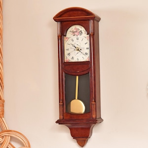 May include: A dark brown wooden wall clock with a rectangular frame, an arched top, and a white clock face featuring black Roman numerals. Above the dial is a floral motif. A gold-colored pendulum hangs behind a glass panel in the lower section.