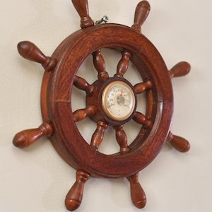 Könnte beinhalten: Ein dunkelbraunes Holzsteuerrad mit acht Speichen und einem kreisförmigen Messgerät in der Mitte. Das Rad hat eine glänzende Oberfläche. Das Messgerät hat ein weißes Zifferblatt mit schwarzen Zahlen und einem Zeiger. Nautische Dekoration.