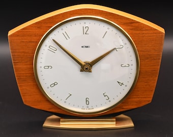May include: A Metamec mantel clock with a wood-grain finish, a white circular face, and gold-toned hands and numerals. The clock features a distinctively shaped wooden frame and a gold-colored metal base, with the brand name Metamec printed on the dial.
