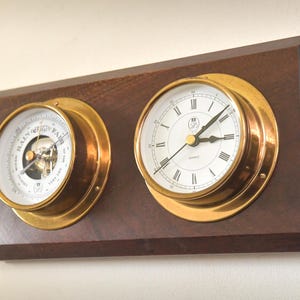 Könnte beinhalten: Eine hölzerne Wetterstation mit Barometer und Uhr. Das Barometer hat ein weißes Zifferblatt mit schwarzer Schrift und eine goldfarbene Lünette. Die Uhr hat ein weißes Zifferblatt mit schwarzen römischen Ziffern und goldenen Zeigern.