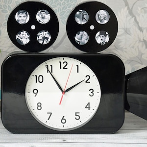 Puede incluir: Un reloj negro con forma de cámara de cine vintage. La esfera blanca del reloj tiene números y manecillas negras. Dos elementos negros con forma de carrete de película en la parte superior muestran fotos en blanco y negro de estrellas de cine clásicas.