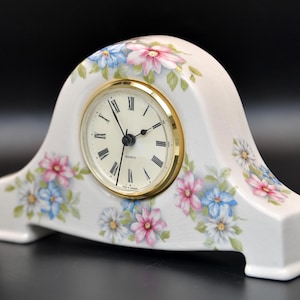 May include: White ceramic mantel clock with a gold-tone clock face and Roman numerals. The clock is decorated with pink, blue, and white floral designs. The clock is approximately 6 inches tall.
