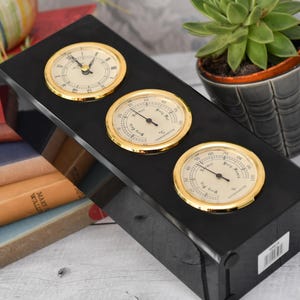 Puede incluir: Estación meteorológica negra con tres esferas doradas: un reloj, un termómetro y un higrómetro. La unidad se encuentra cerca de libros y una suculenta en maceta. Los diales miden la temperatura en grados Celsius.