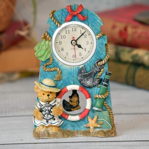 May include: A nautical-themed quartz clock with a light blue wooden frame. The clock features a teddy bear in a sailor outfit, an anchor, a life preserver, and a starfish. The clock face has black numbers and hands.