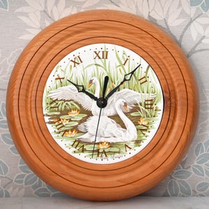 May include: A round wall clock with a polished light brown wooden frame. The clock face features a white background with a painted illustration of two white swans swimming in a pond with green reeds and orange water lilies, and Roman numerals.