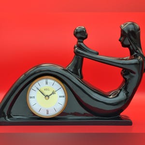 May include: A glossy black ceramic mantel clock featuring a stylized figure of an adult holding a small child. The clock face is circular with a pale yellow center, black hands, and Roman numerals. The text WBL is printed on the clock face.