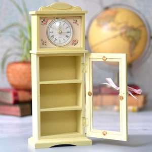 May include: A miniature pale yellow wooden clock and cabinet. The clock face has Roman numerals and floral details. The cabinet has three shelves and a glass door, which is ajar. A pink ribbon is tied to the door. The clock is a decorative item.