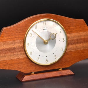 May include: A mantel clock with a horizontal wooden body featuring a natural grain pattern. The circular clock face has a gold-toned bezel, a silver centre, and gold-coloured numerals. It stands on a rectangular wooden base with small gold feet.
