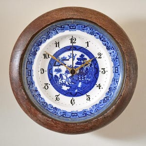 May include: Round wall clock with a brown wooden frame. The clock face is a white plate with a blue and white Asian-inspired design. Gold-colored hands point to the time. The clock is a decorative item.
