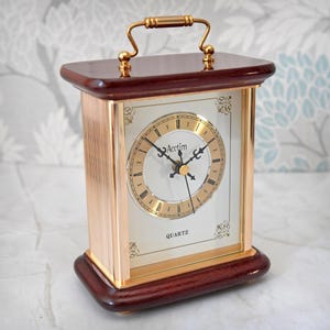 May include: A rectangular, vintage-style clock with a gold-tone face and Roman numerals. The clock has a dark brown wooden base and top, with gold-colored metal accents and a handle. The clock face reads "Aectim Quartz".