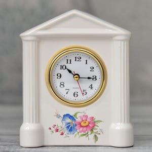 Può includere: Un orologio in ceramica bianca con una lunetta dorata e un motivo floreale. L'orologio ha una forma architettonica classica con un frontone e colonne scanalate. Il quadrante mostra numeri e lancette nere, con una lancetta dei secondi rossa.