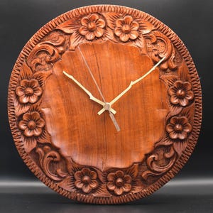 May include: A round wooden wall clock featuring intricate hand-carved floral patterns and scrolling leaf designs along the outer border. The centre of the clock is smooth with a natural wood grain, equipped with three thin, light-coloured metal hands.