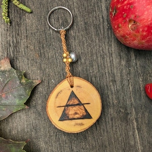May include: A wooden keychain with a silver key ring. The round wooden pendant features a dark triangle design with a horizontal line. The keychain is attached to a woven cord with gold beads and a silver bead. The background is a wooden surface.