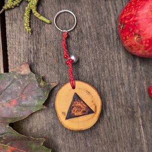 May include: A wooden keychain with a circular wooden pendant featuring a dark triangle design. The keychain has a red cord with small red beads and a silver bell. The background includes leaves and a red fruit on a wooden surface.