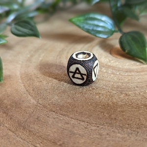 May include: A small, wooden bead with a dark brown, textured exterior. The bead features carved white symbols on its sides, including an upward-pointing triangle within a circle. The bead is displayed on a light brown, wooden surface, with green leaves in the background.