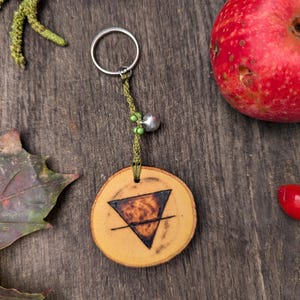 May include: A wooden keychain with a green cord and a silver charm. The wood is light brown and has a dark brown triangle with a line across it. The keychain is on a wooden surface with a red apple and green leaves.