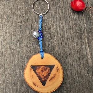 May include: A wooden keychain with a blue cord and a silver bell. The wood is a light brown color and has a dark brown triangle burned into it.