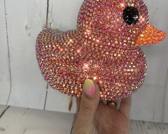 May include: A pink rubber ducky covered in iridescent rhinestones. The duck has a black eye and an orange beak.
