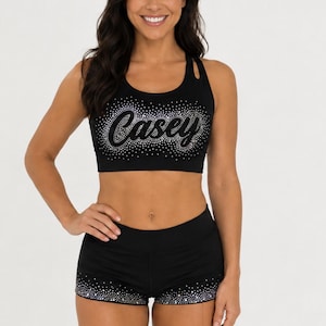 May include: A black athletic crop top and matching shorts set. The crop top features a racerback design and the name Casey in script font, surrounded by silver rhinestones. The shorts have a matching silver rhinestone trim along the leg openings.